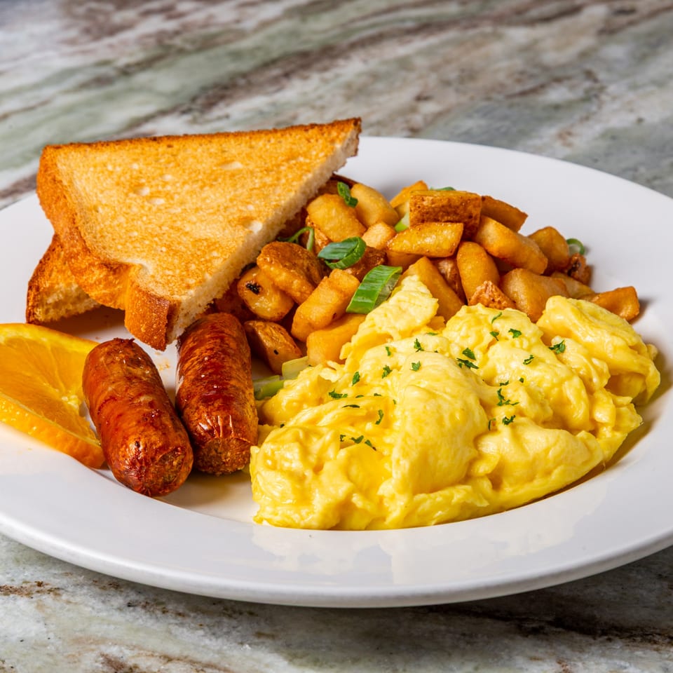 A Big John's Eatery breakfast plate with scrambled eggs, sausage links, home fries, toast, and an orange slice.