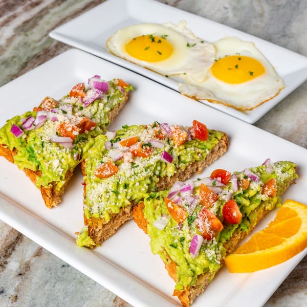 Avocado toast with tomatoes, red onion, and cheese, served with two fried eggs and an orange slice.