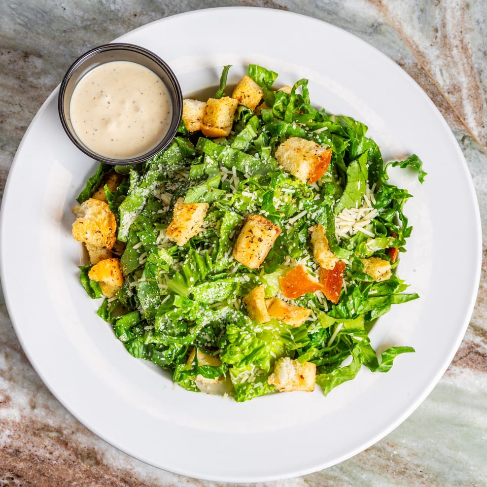 Overhead view of a Caesar salad with croutons, parmesan, and a side of dressing on a white plate.