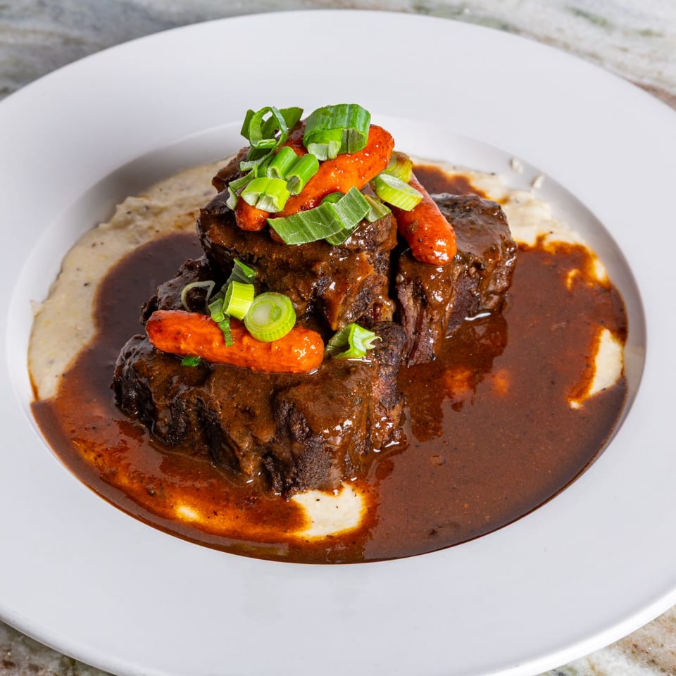 Braised short ribs with gravy, carrots, and green onions, served over grits on a white plate.